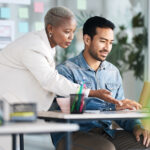 An example of a mentoring relationship, a black woman with short white hair assists a latino man at his laptop.